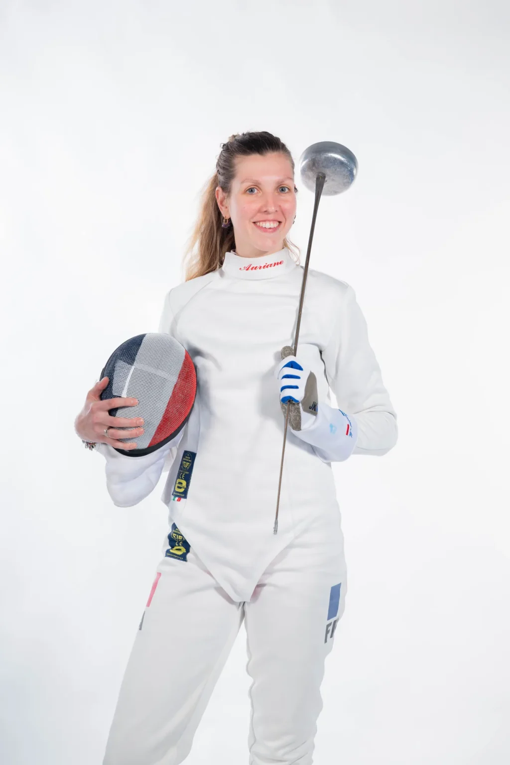 Auriane Mallo souriante, masque sous le bras et épée dressée, sur fond blanc