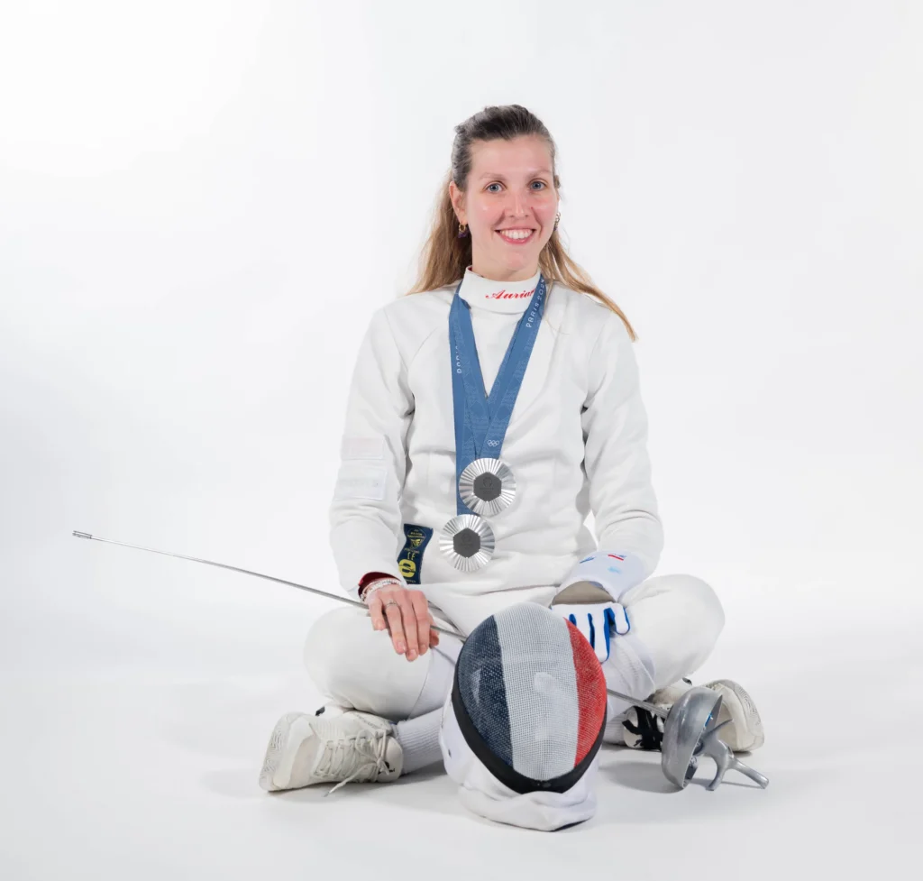 Auriane Mallo assise en tailleur, masque posé devant elle et médailles autour du cou, sur fond blanc