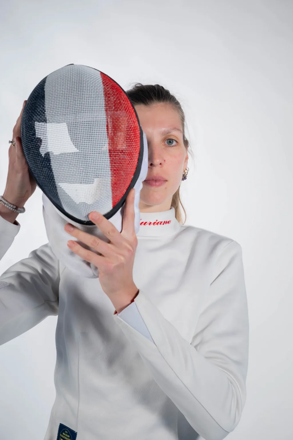 Auriane Mallo cachant son visage derrière son masque d'escrime, sur fond blanc