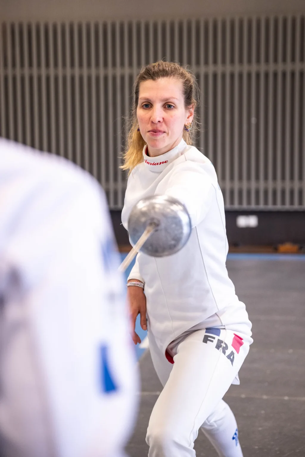 Auriane Mallo concentrée, épée devant elle, face à une adversaire floue au premier plan