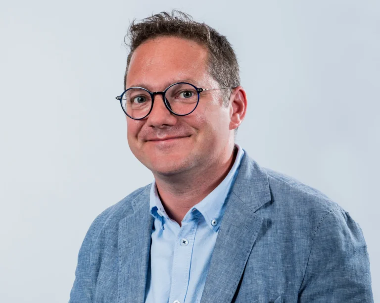 Portrait corporate en studio d'un membre de l'équipe Yabuko en blazer bleu clair et lunettes, souriant, sur fond clair